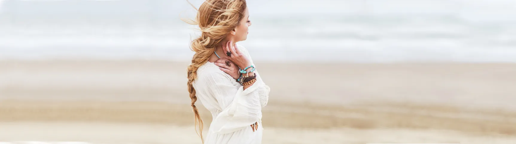 Verträumte hübsche junge Frau am Strand. Sie ist sommerlich bekleidet und schaut zur Seite. Am Hals und am Handgelenk trägt sie bunten Modeschmuck.